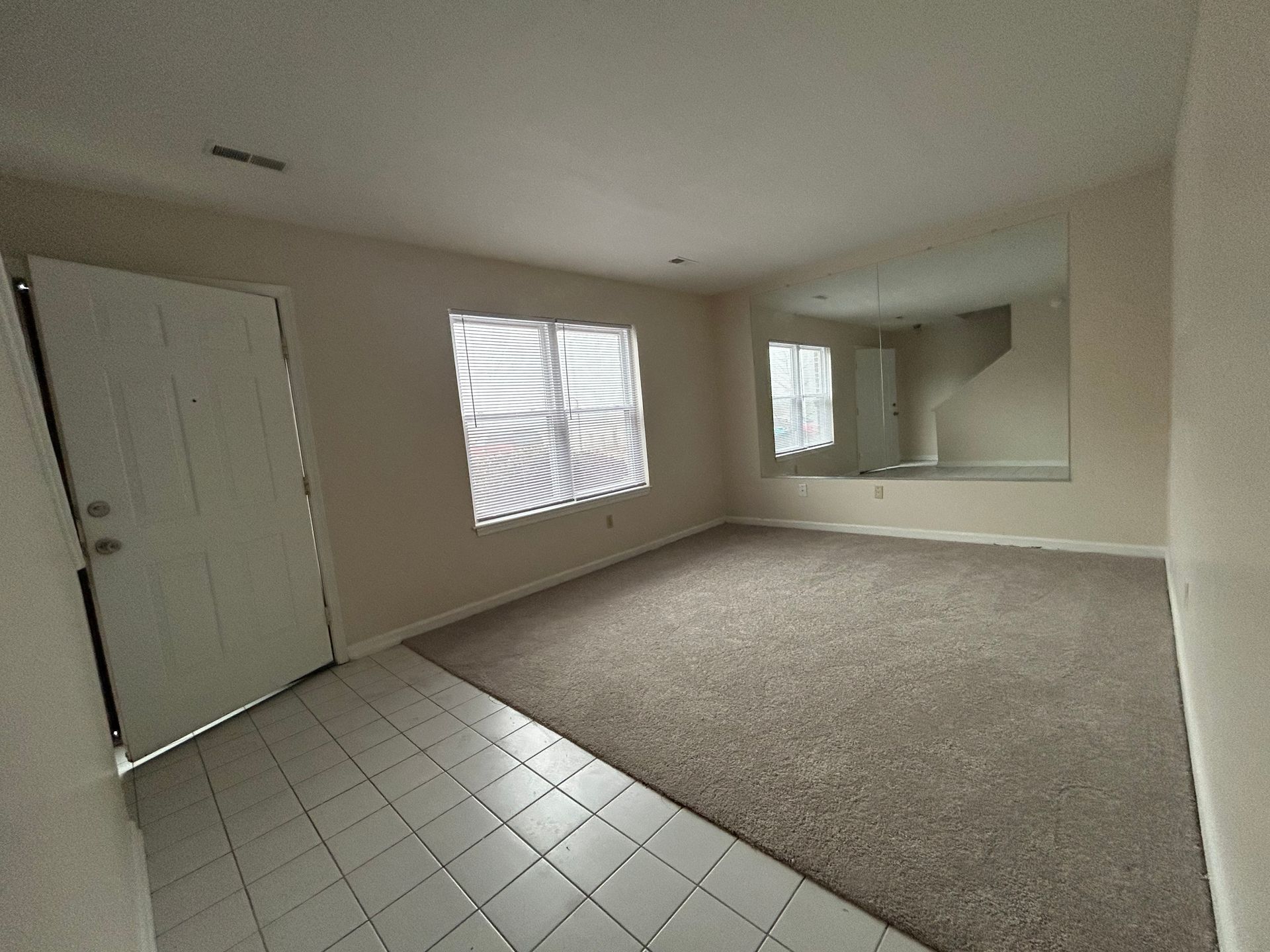An interior view of a room with a tiled entryway, tan carpeted flooring, a white front door, and a mirrored wall.