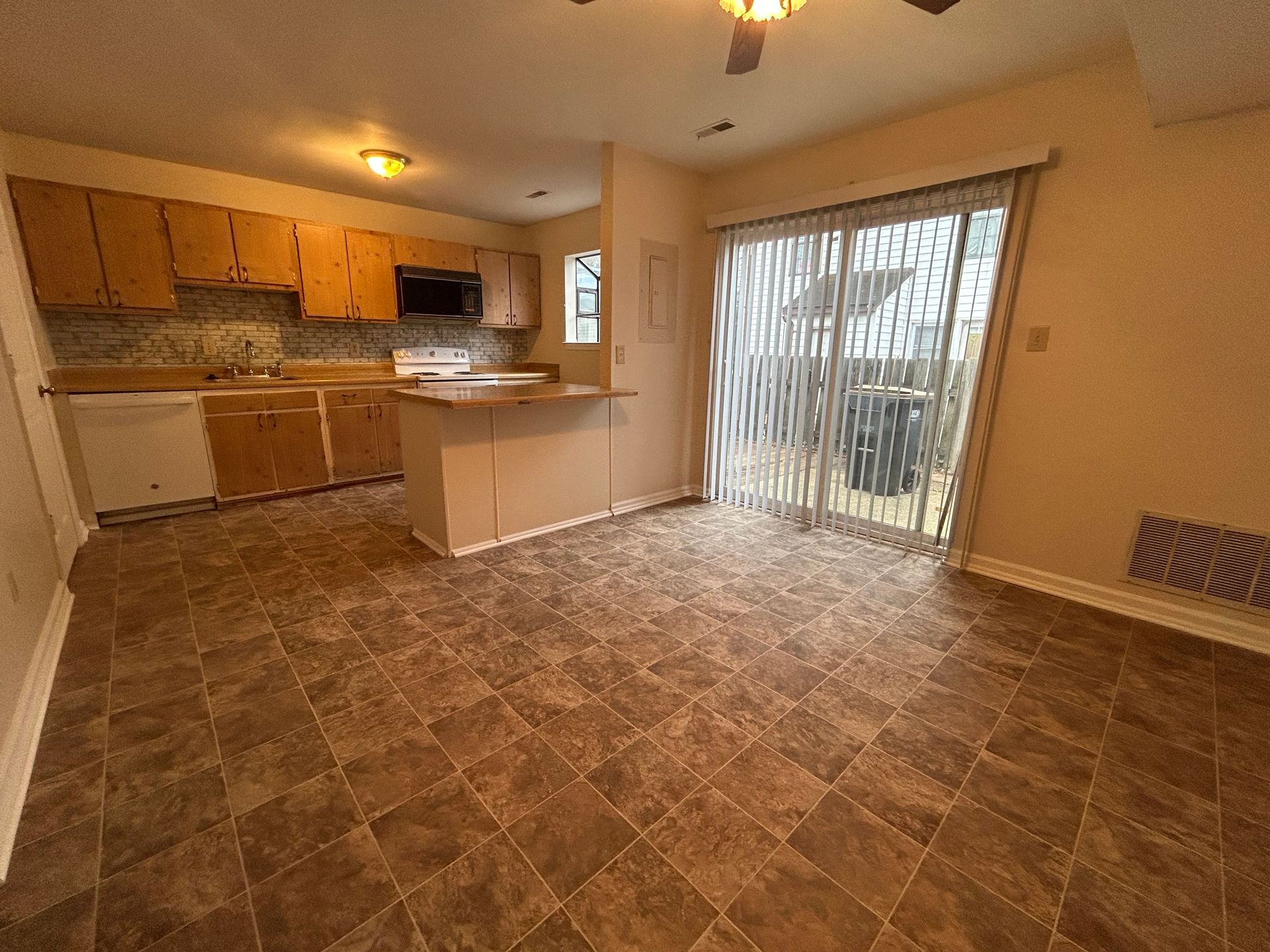 An open-plan kitchen and dining area with light wood cabinets, dark countertops, brown patterned flooring, and sliding door.