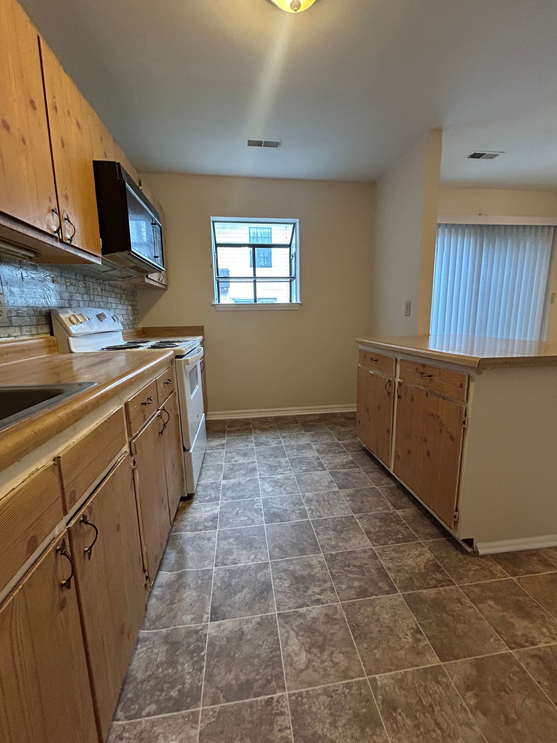 A kitchen with light wood cabinets, brown patterned linoleum flooring, a window, and an adjacent room with blinds.