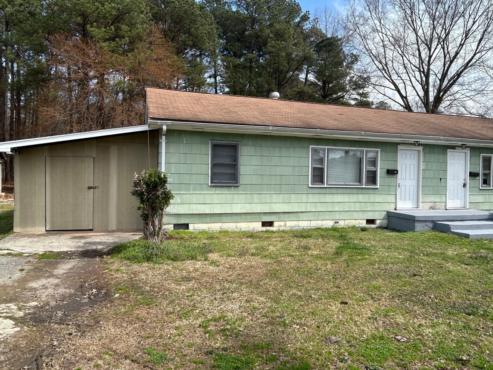 A single-story, light green rectangular house with a brown shingled roof, two front doors, and a small attached shed.