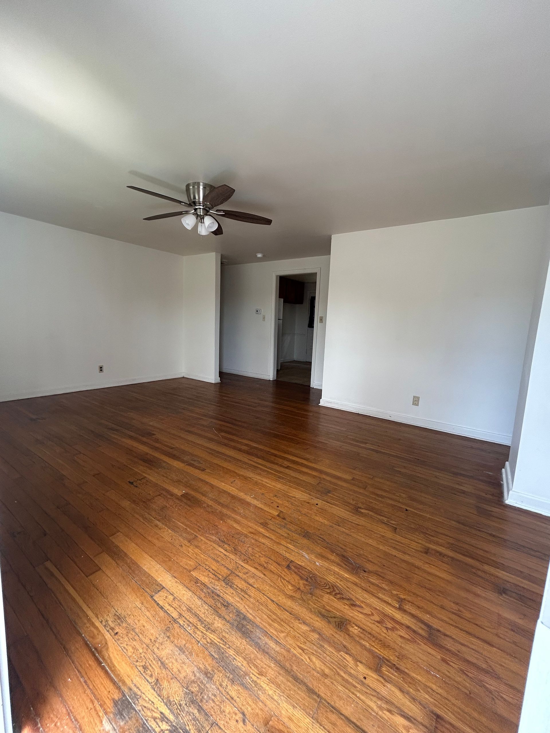 An empty room with wood flooring, white walls, and a ceiling fan, looking toward a doorway leading to another area.
