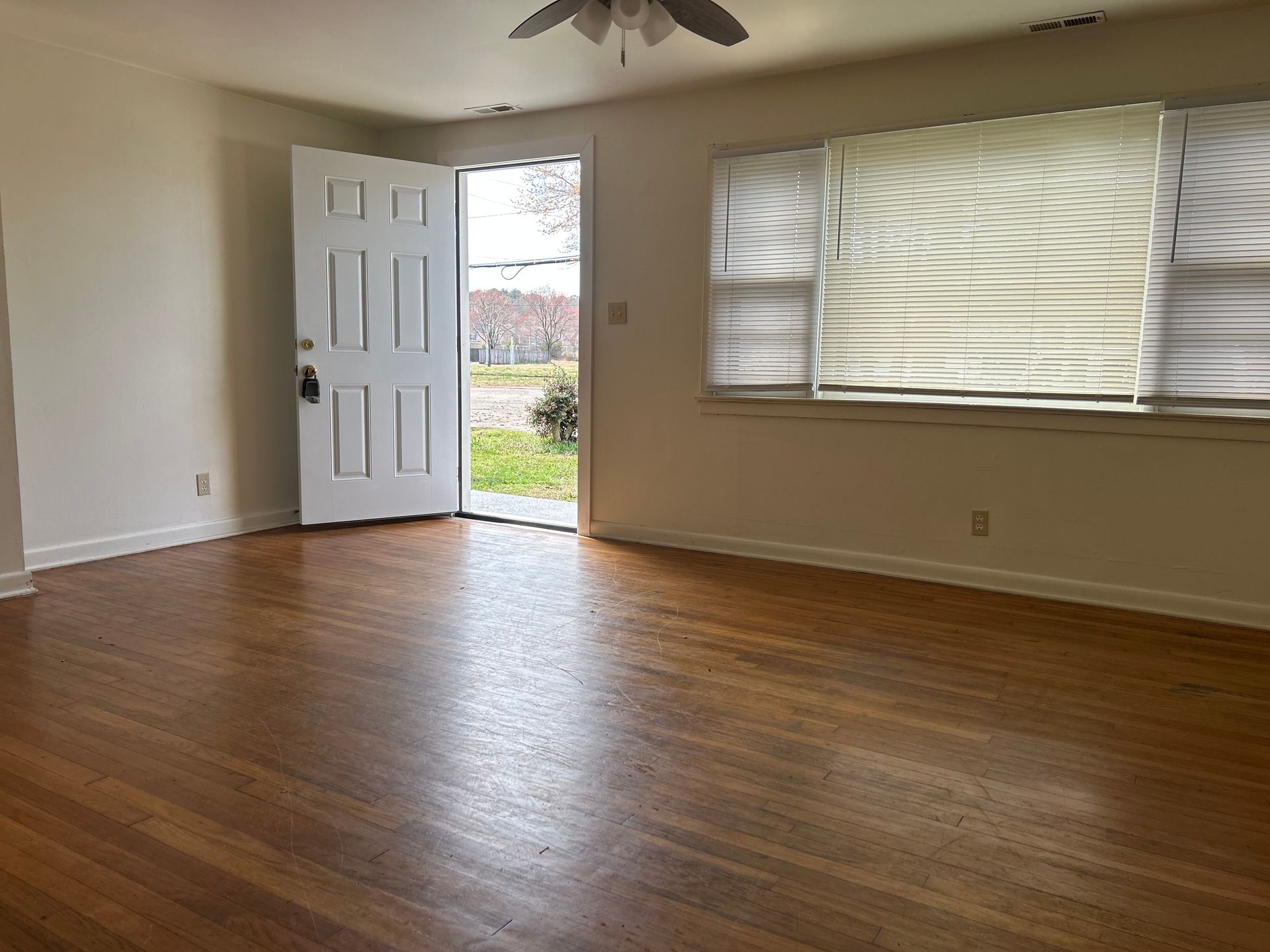 A bright, empty room with light wood floors, white walls, a white front door left open, and a large window with blinds.