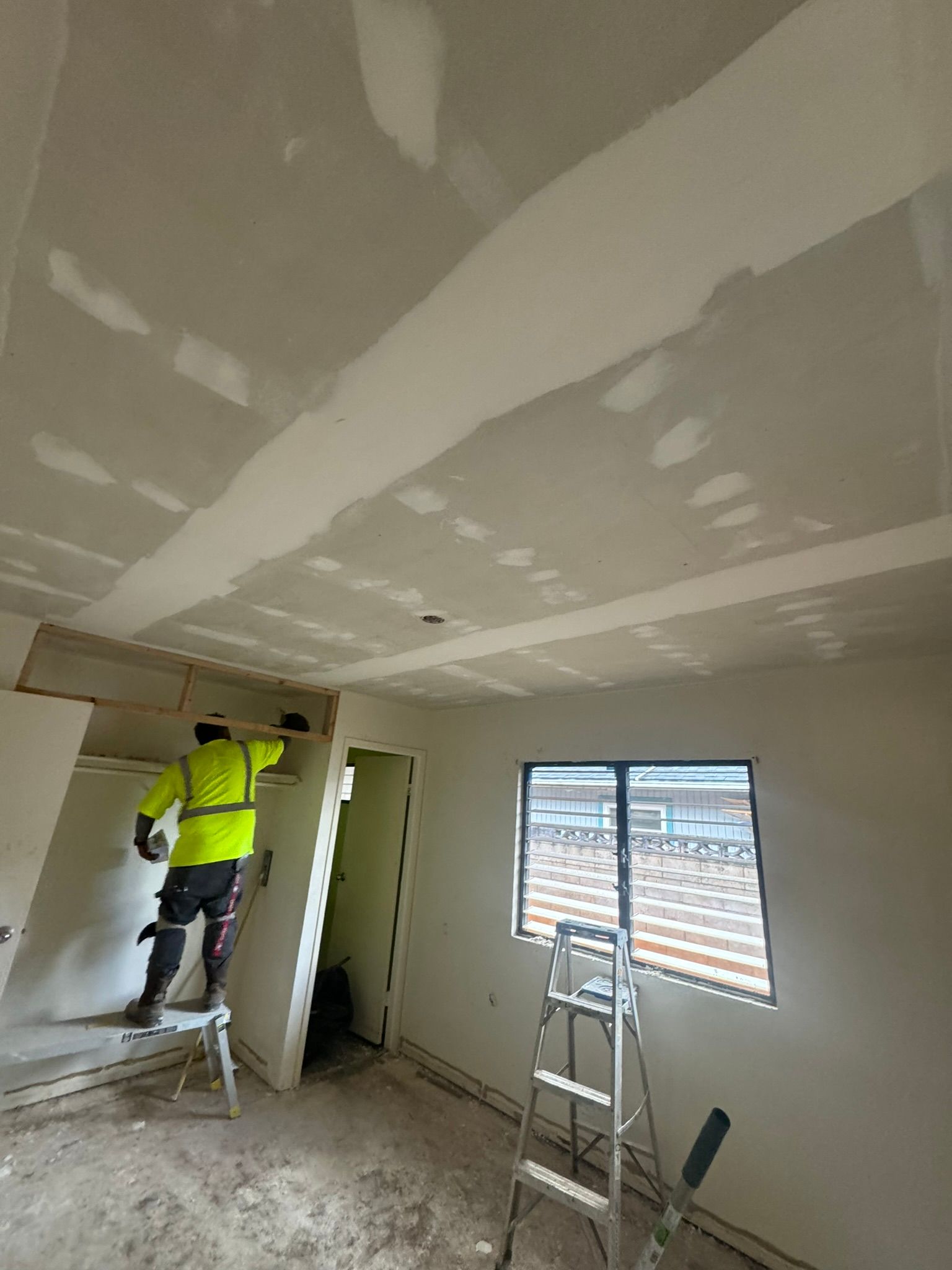 A construction worker in a safety vest working on drywall on a ceiling indoors.