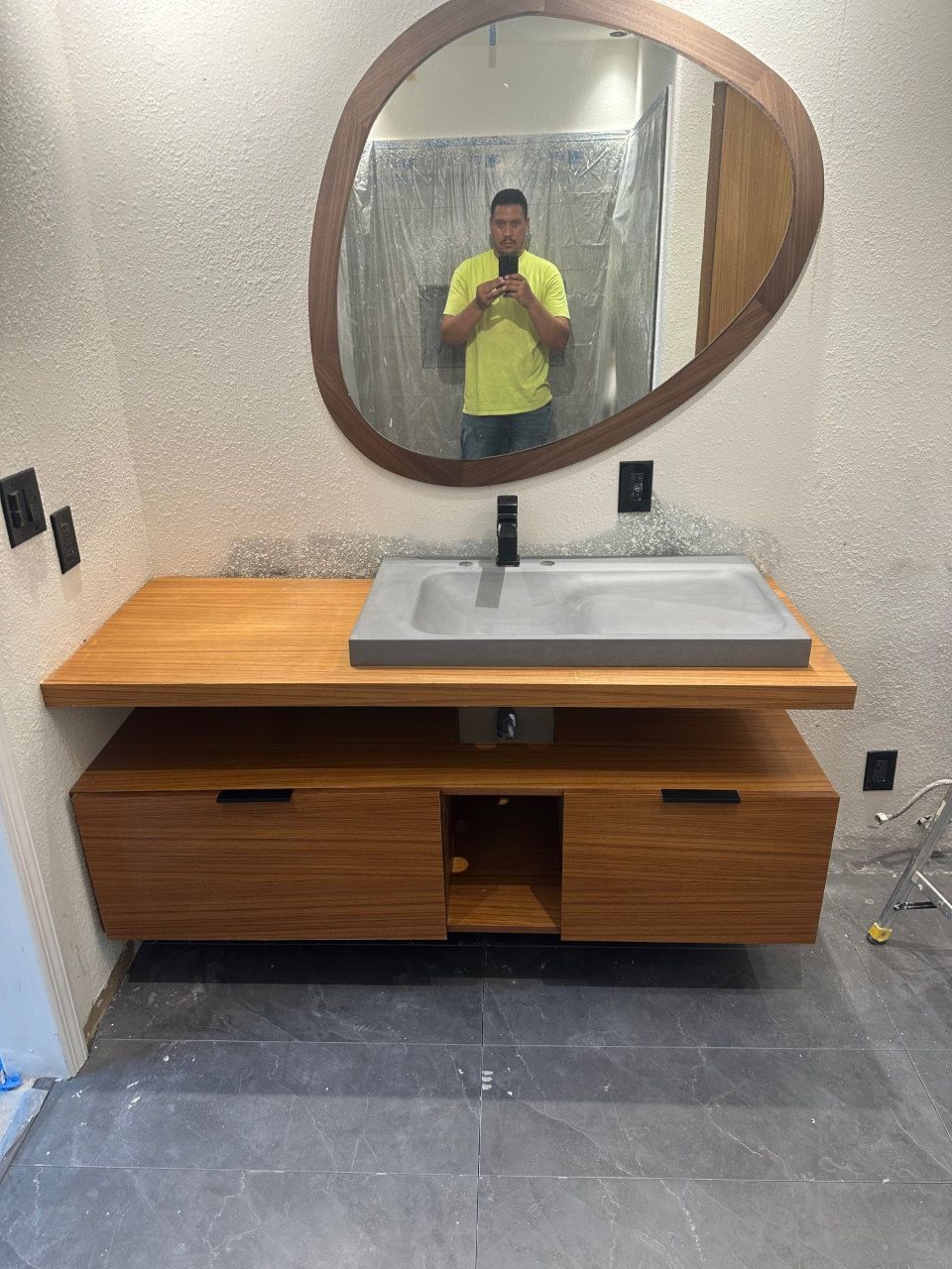 Bathroom vanity with wood cabinet, grey sink, and oval mirror; person taking photo in reflection.