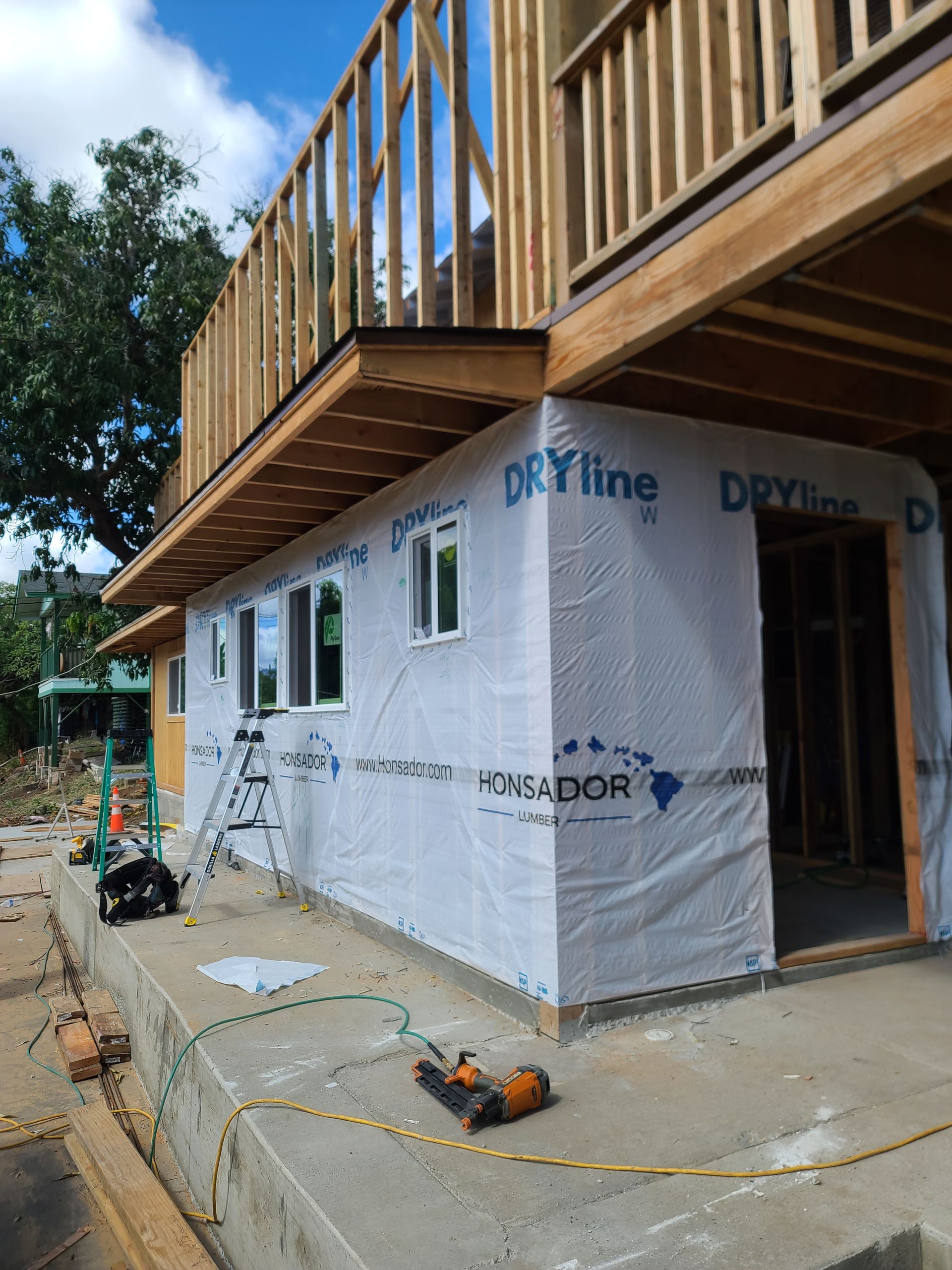 Building exterior under construction, covered in blue waterproof wrap. Two-story structure, wooden framing, gray concrete base.
