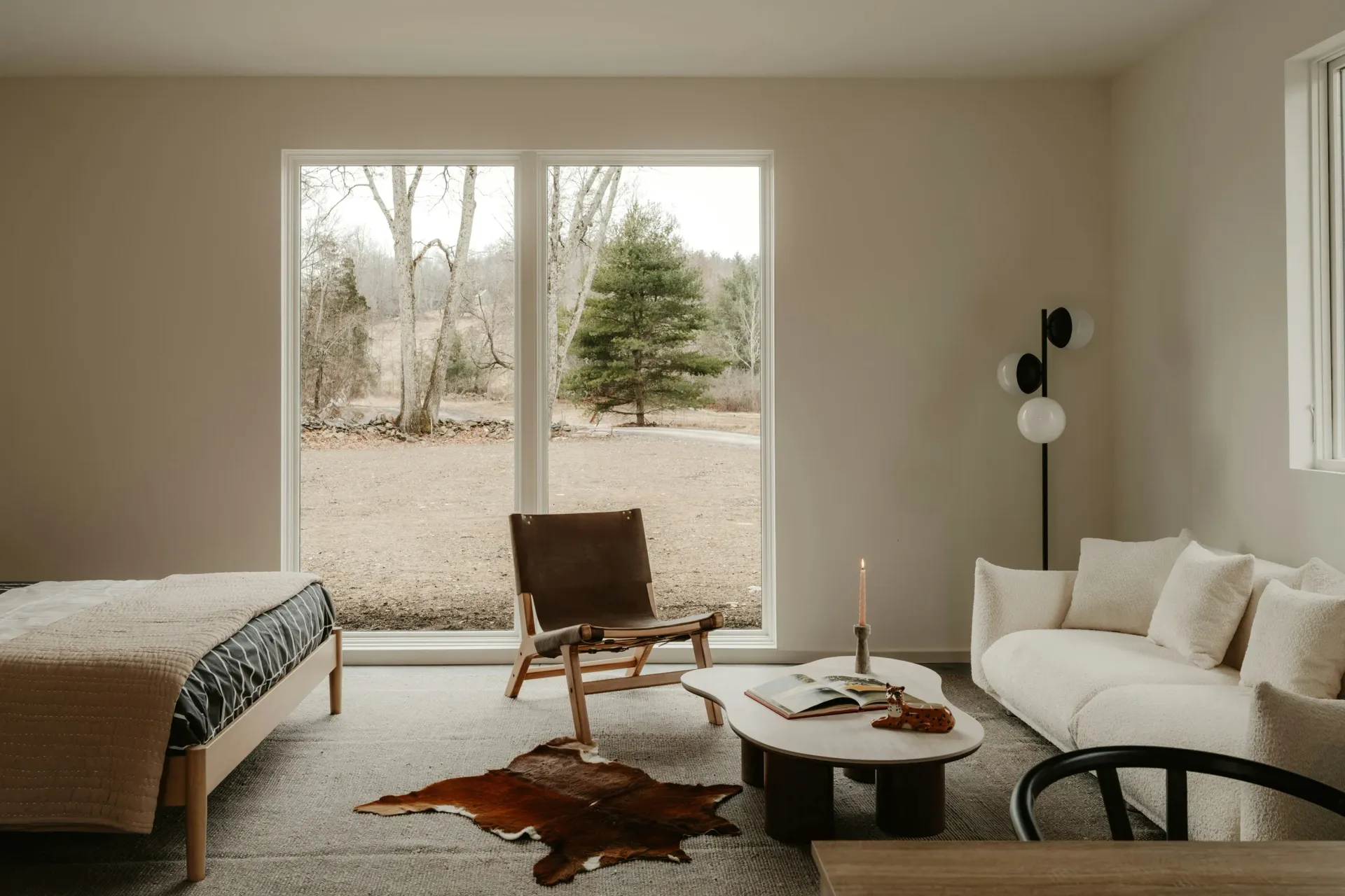A modern, minimalist living room with a white sofa, patterned lounge chair, coffee table, and large window to a wood view.