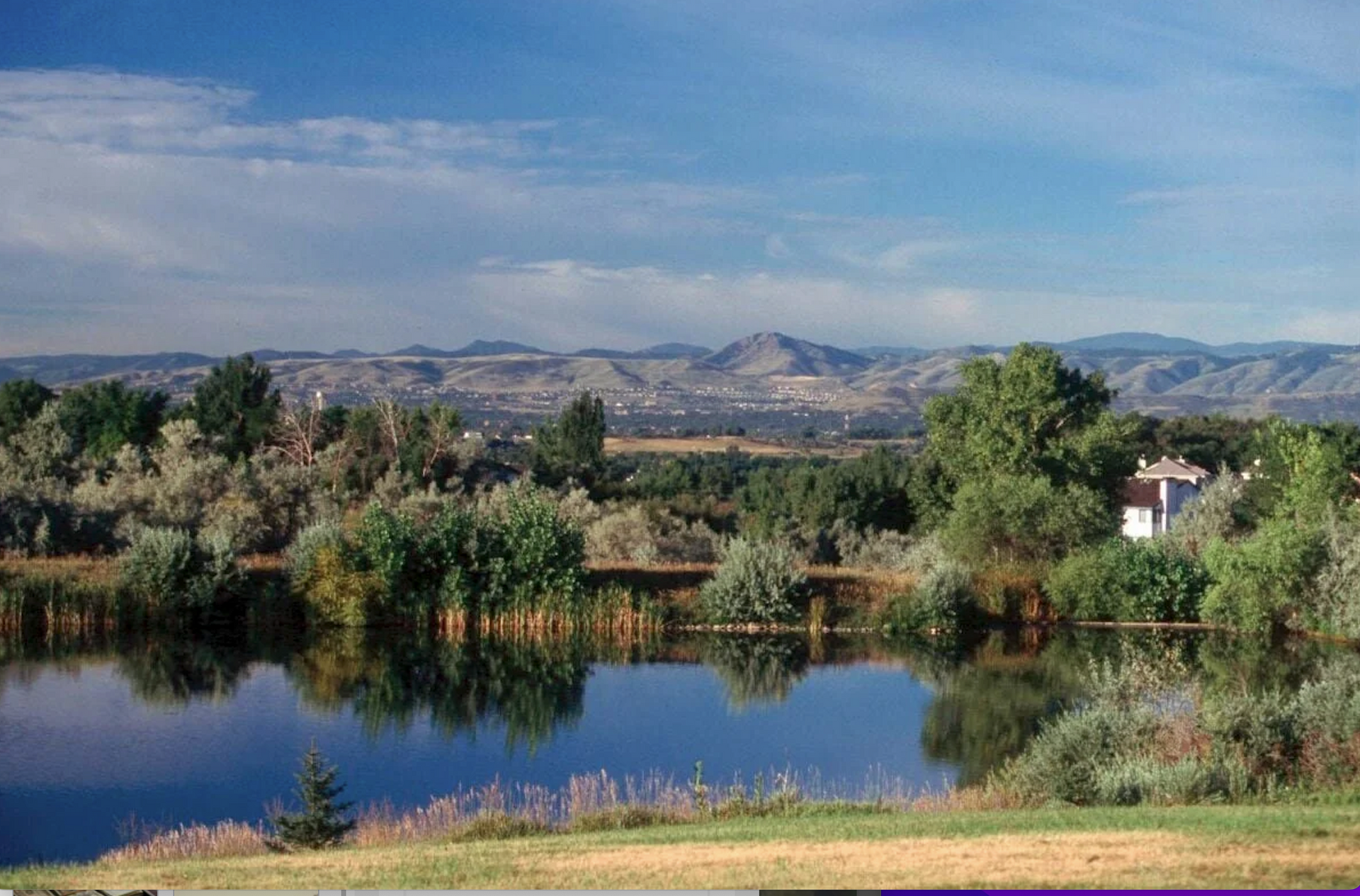Calm lake reflects trees and sky in Arvada CO; distant mountains under a blue sky.