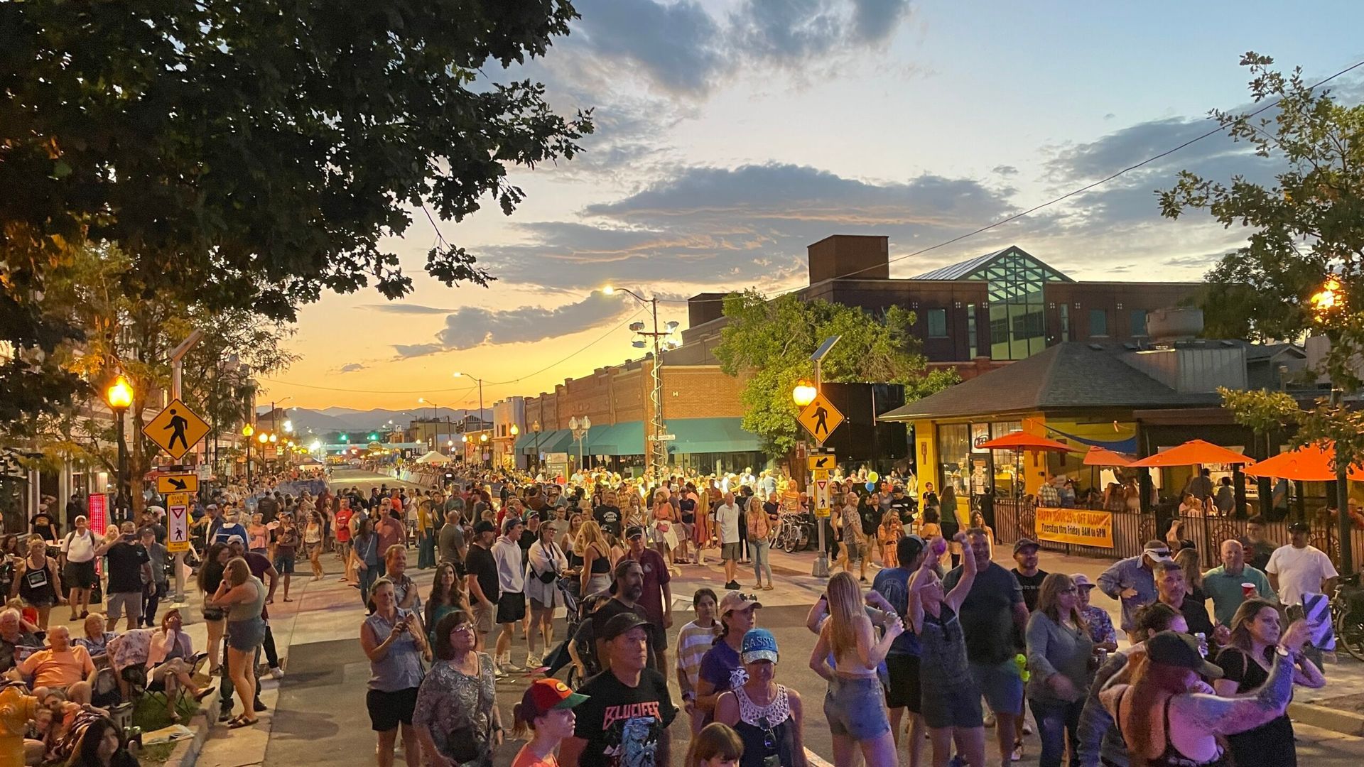 Large crowd on a street at sunset with shops, trees, and buildings. People are gathered, with some looking up.