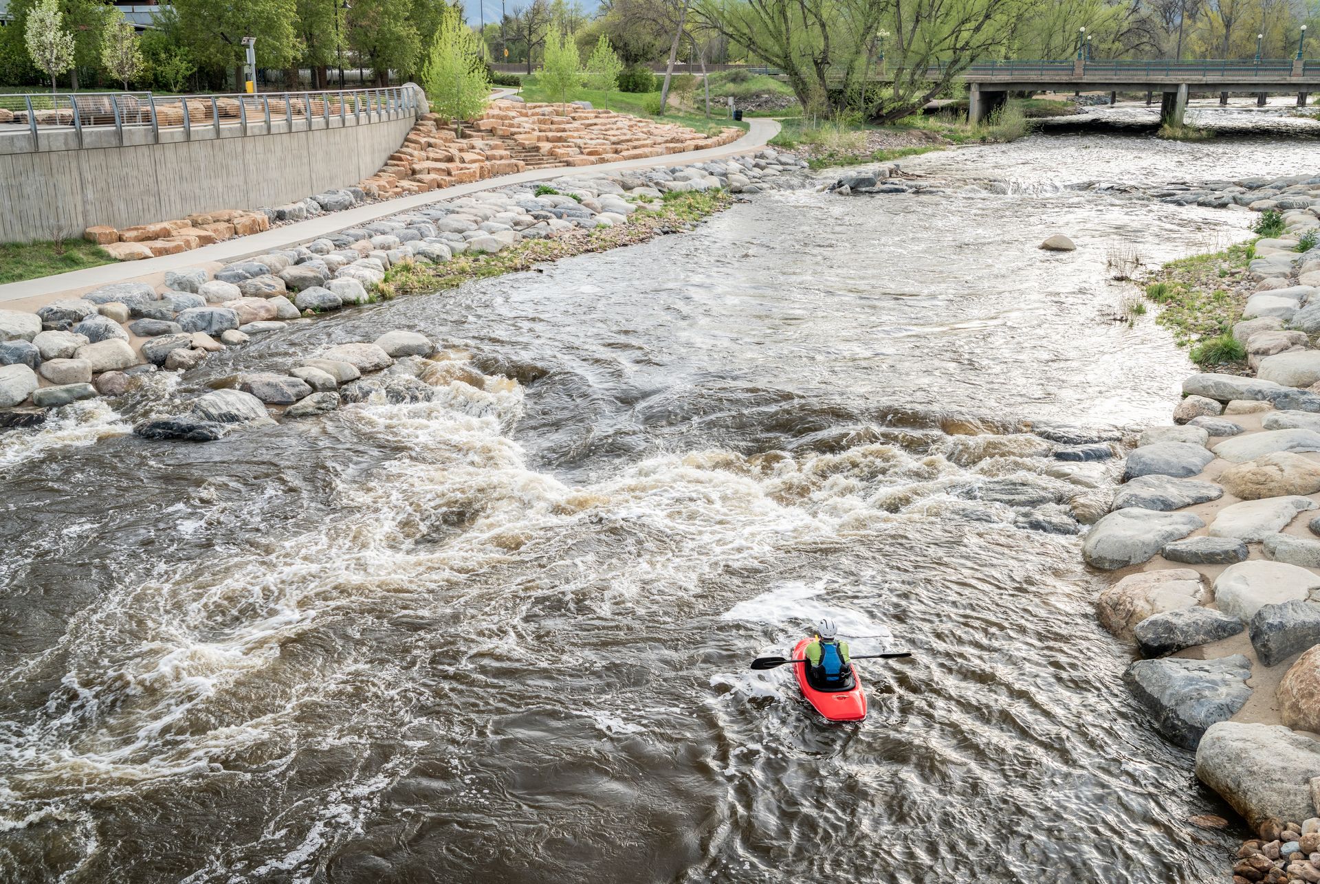 A small red boat floats in a fast-moving, rocky river near a stone embankment.
