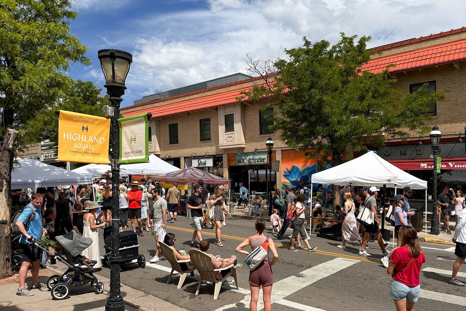 Street fair with crowds, tents, shops, and people relaxing on chairs, in a sunny setting.