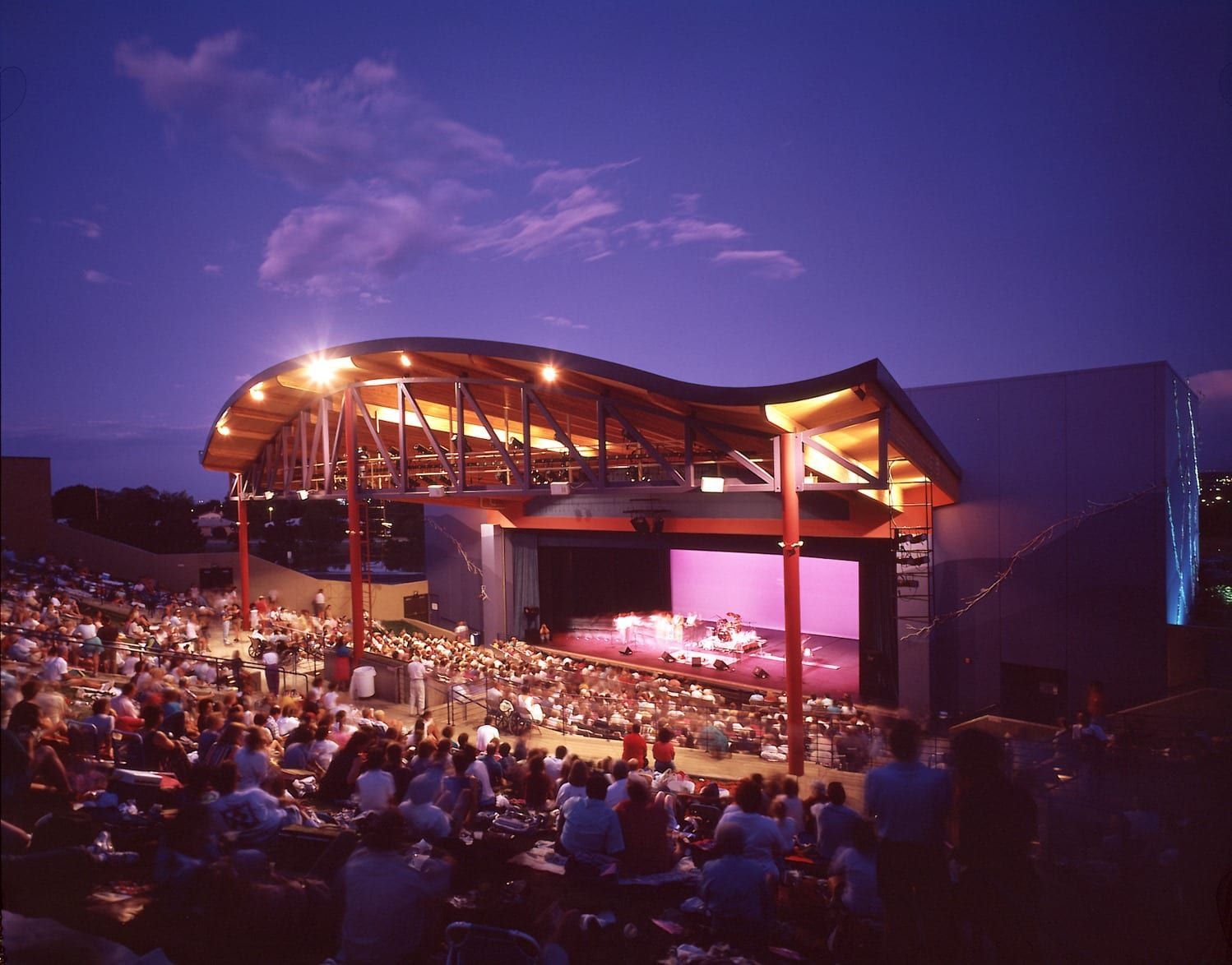 Outdoor theater with stage and audience at dusk. Curved wooden roof, purple sky.