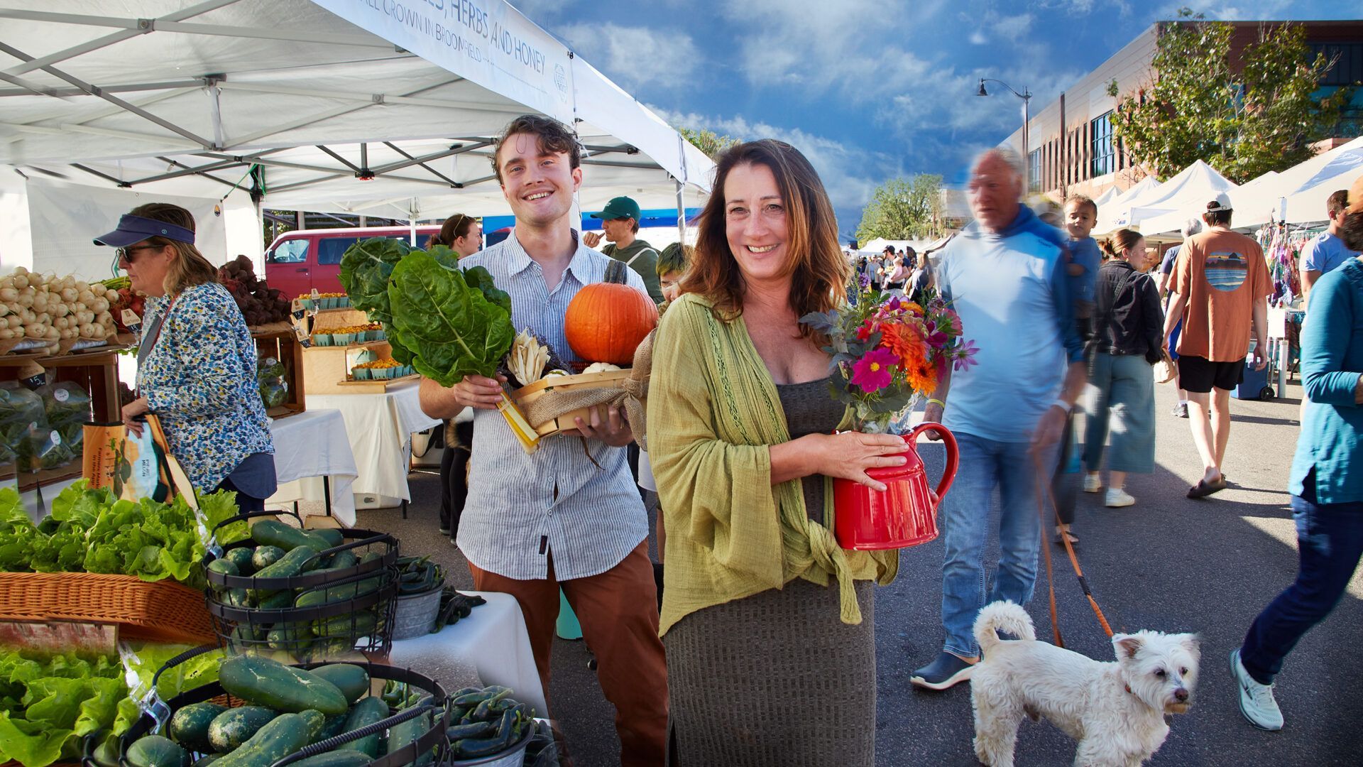 People at a farmers market, holding produce and flowers. White tents and a mountain backdrop. Sunny day.