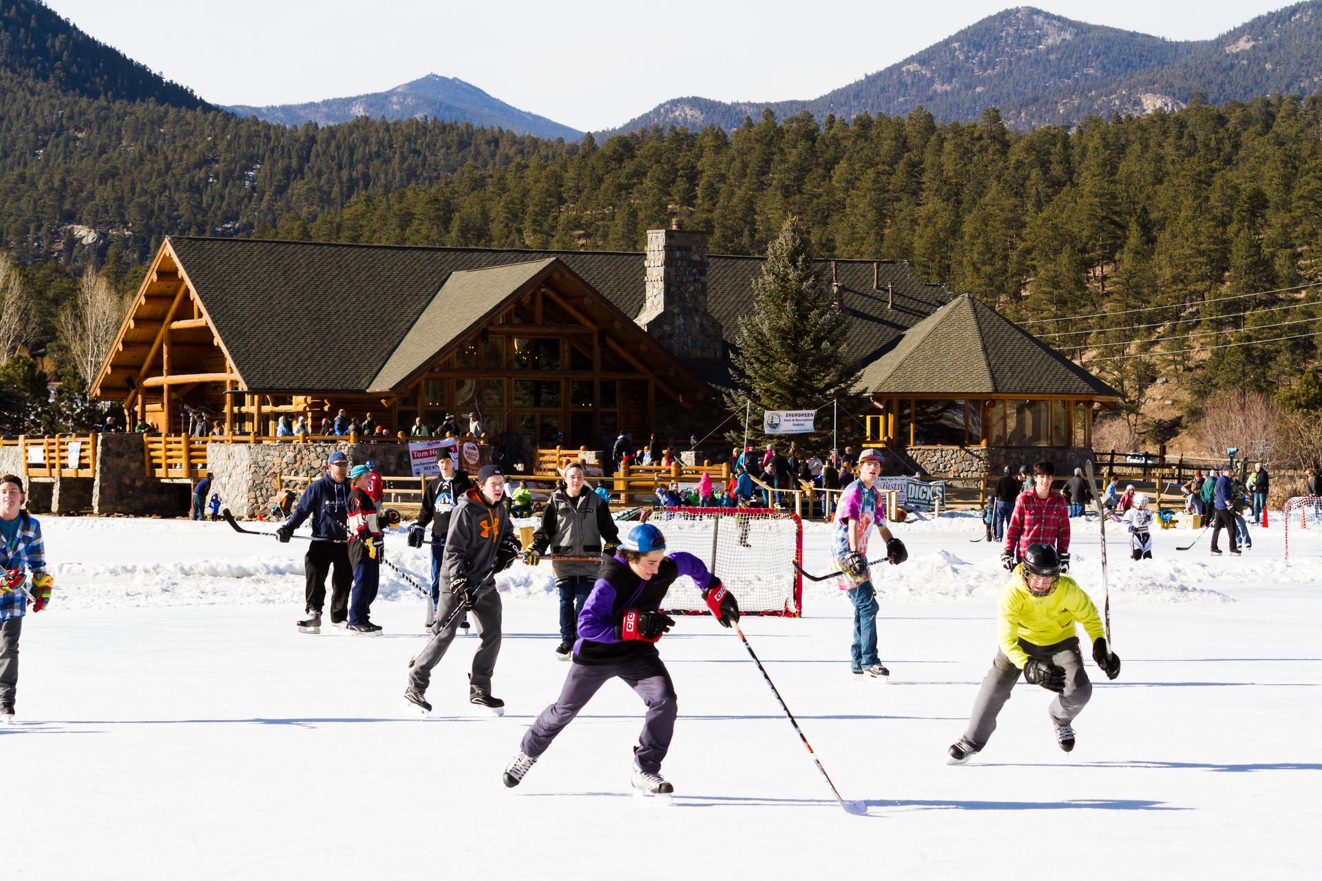 People playing ice hockey on a frozen lake in front of a lodge, mountains in the background.