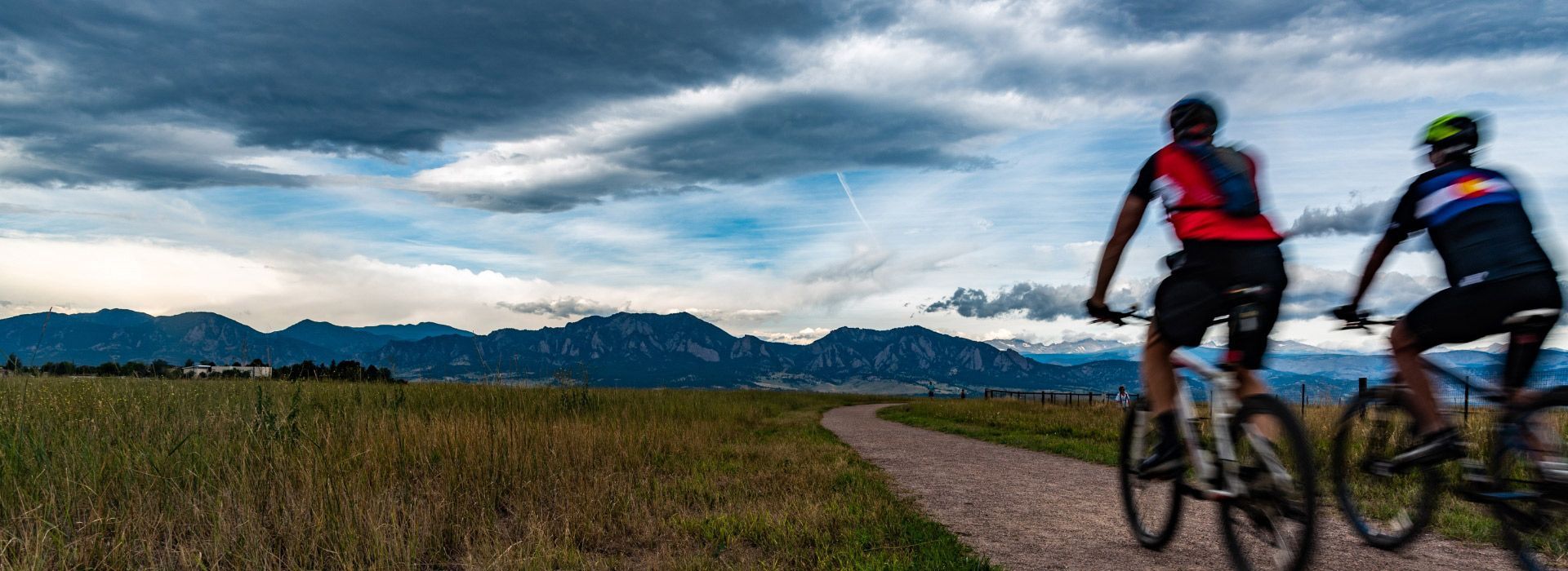 Two cyclists on a path, riding toward mountains under a cloudy sky.