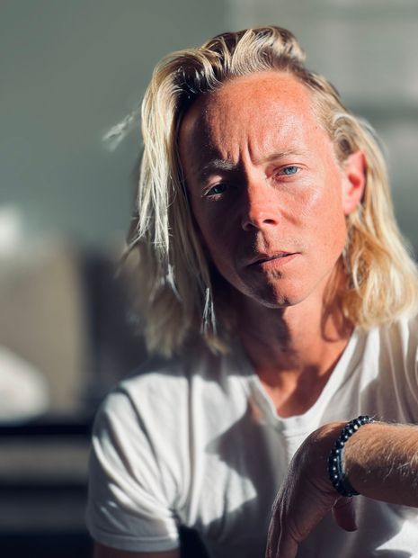 A man with long blonde hair is wearing a white shirt and a bracelet.