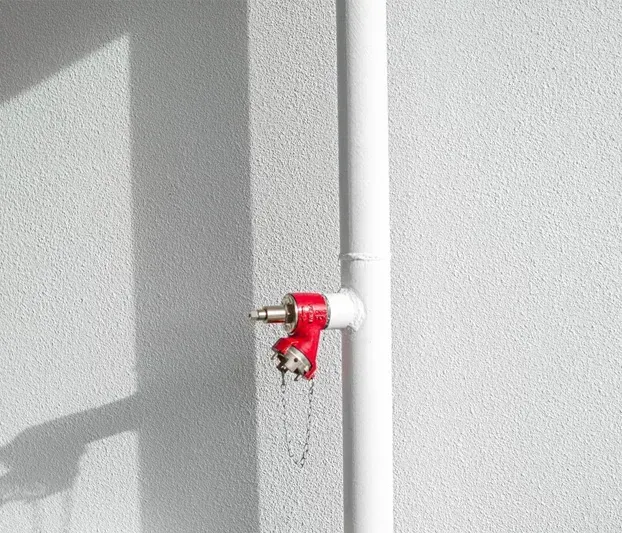 A red valve is attached to a white pipe against a white wall.