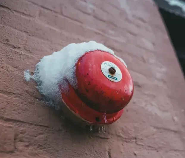 A red fire alarm is covered in snow on a brick wall.