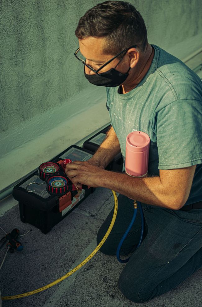 Technician kneeling by HVAC gauges, checking a unit with a yellow hose and pink insulation on the pipe.