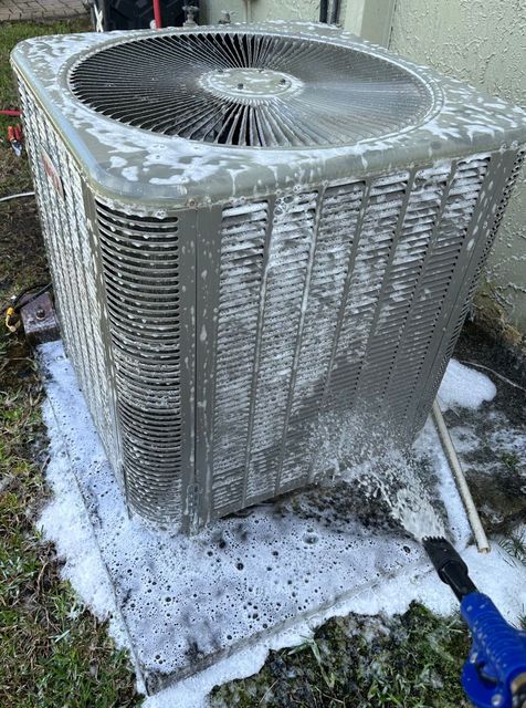 Care to Cool LLC - A person cleaning an outdoor air conditioning unit with soapy water. The unit is covered in foam.