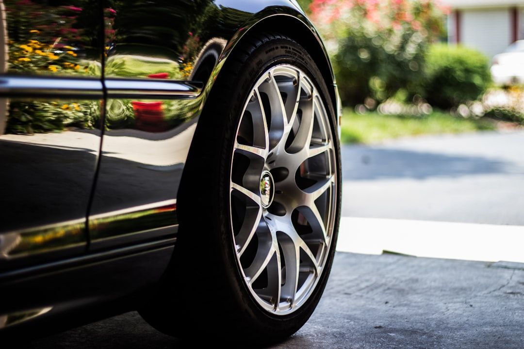 Black car's front wheel, silver rim, parked on gray concrete, with greenery in the background.