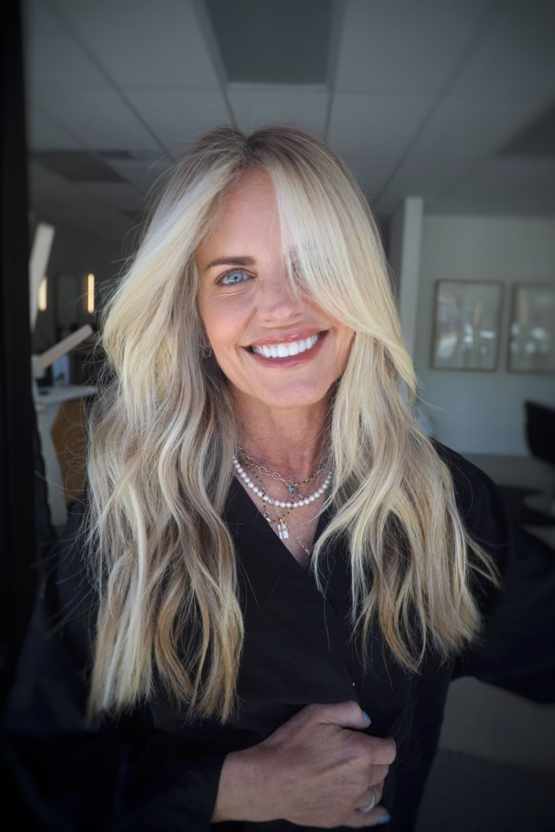 A woman with long blonde hair is smiling and wearing a pearl necklace.