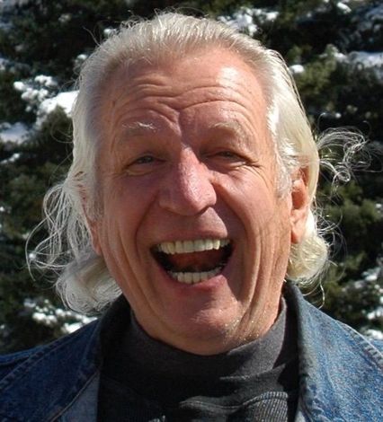 A man with white hair is smiling and wearing a black turtleneck