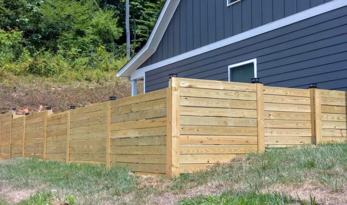 Wooden horizontal plank fence in front of a blue-gray house on a grassy hill.