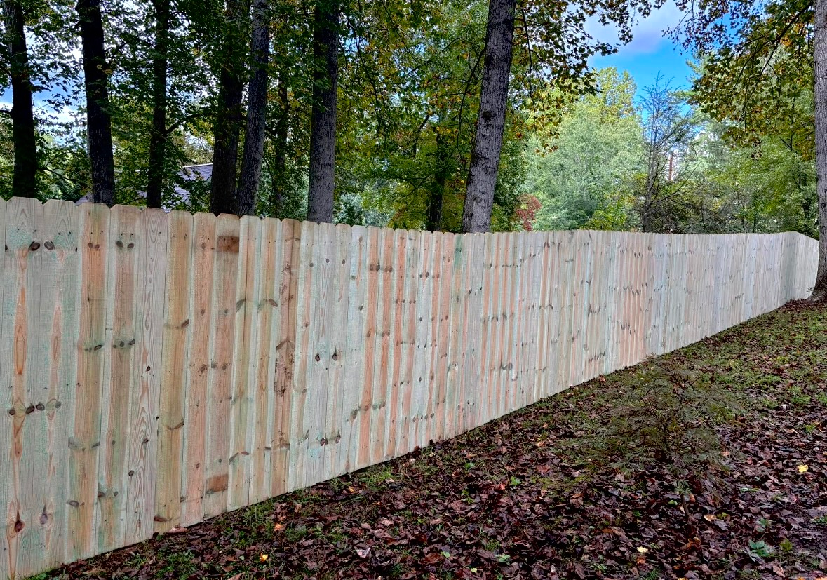 Wooden fence in a wooded area; vertical planks, varying shades of wood, autumn leaves on the ground, trees in the background.
