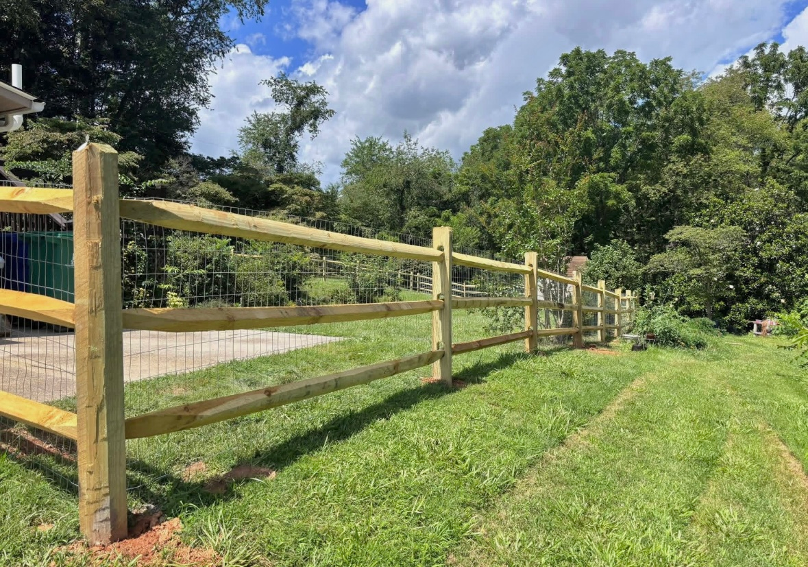 Wooden split-rail fence with wire mesh in a grassy yard, trees in the background, blue sky with clouds.