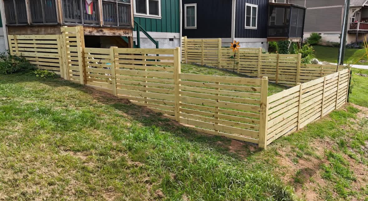 Wooden horizontal slat fence surrounding a grassy yard near houses.