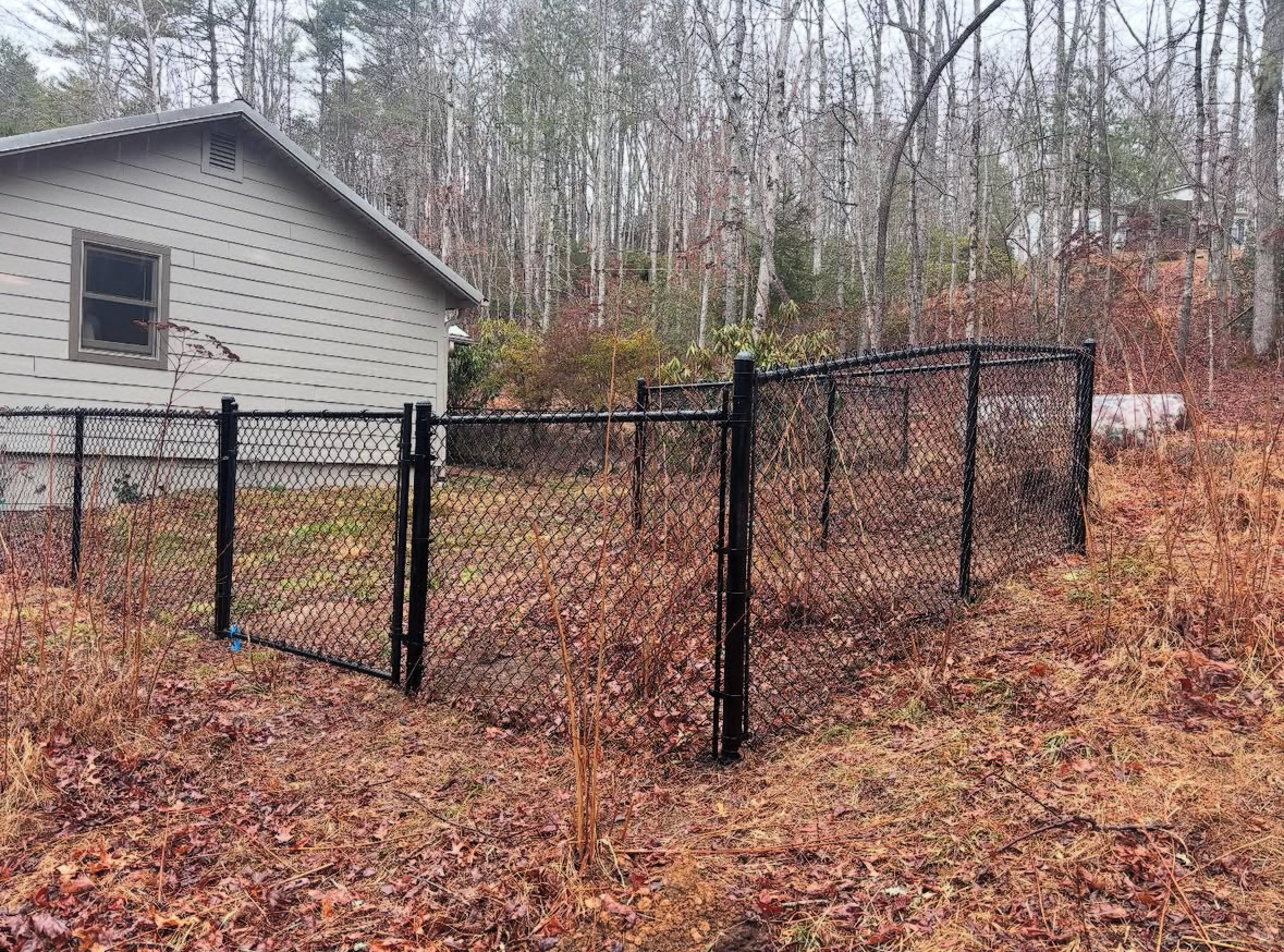 Black chain-link fence surrounds yard next to a tan house; wooded area in the background with brown foliage.