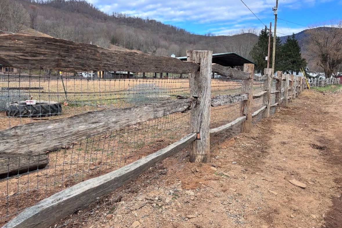Wooden fence with wire mesh, dirt path, rural setting with hills and a barn.