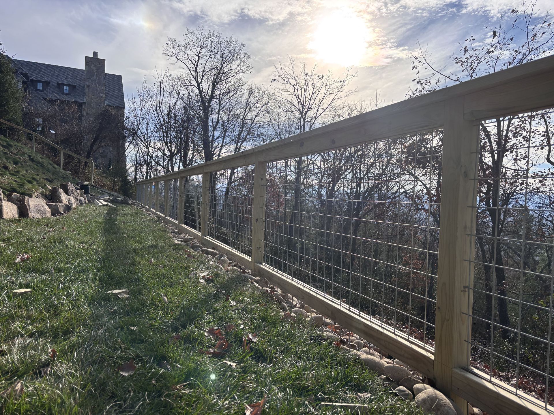 Wooden fence with wire mesh, on a grassy hillside, with a building and trees in the background.