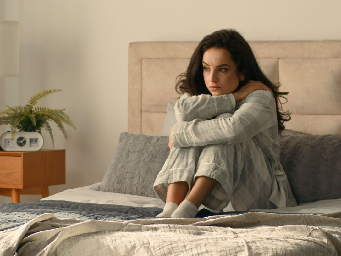 Woman in pajamas sits on bed, hugging her knees, looking sad in a bedroom.