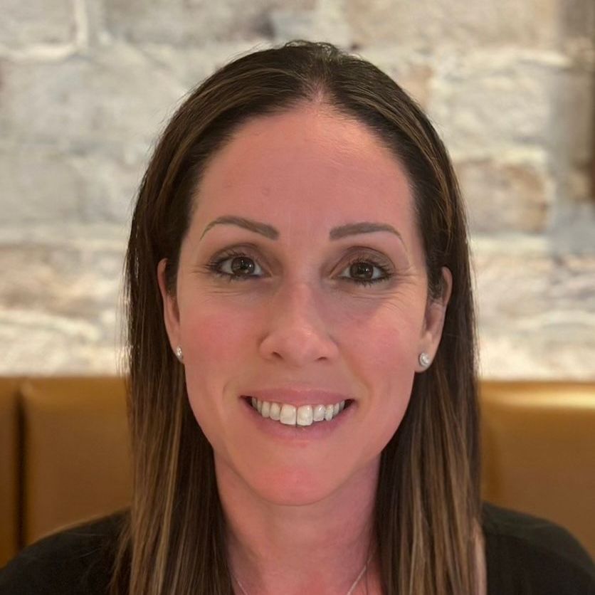 Woman with brown hair smiling, wearing a black top, set against a light-colored stone wall.