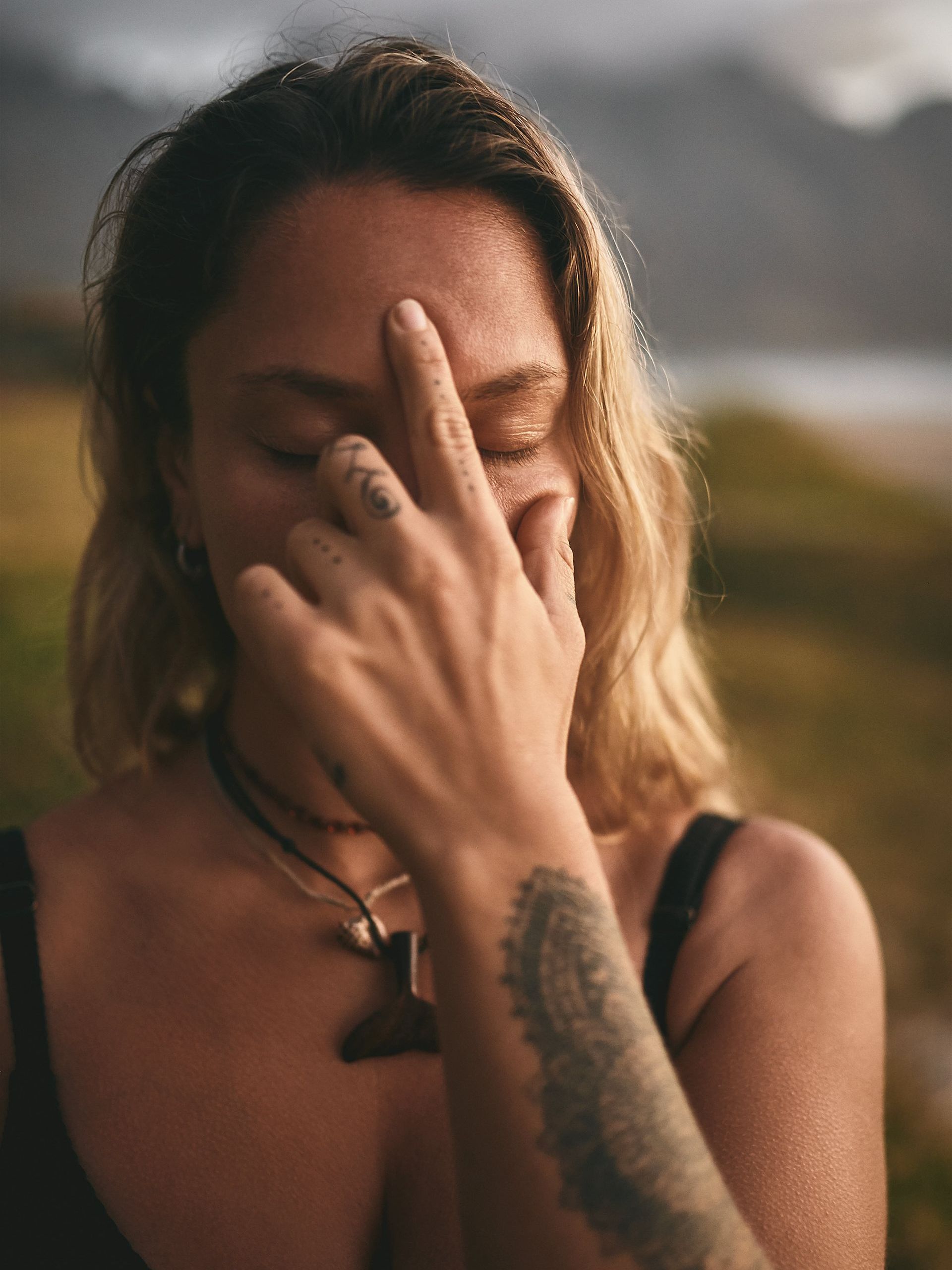Woman with closed eyes, touching forehead with fingers, wearing necklaces and a tattoo on arm.