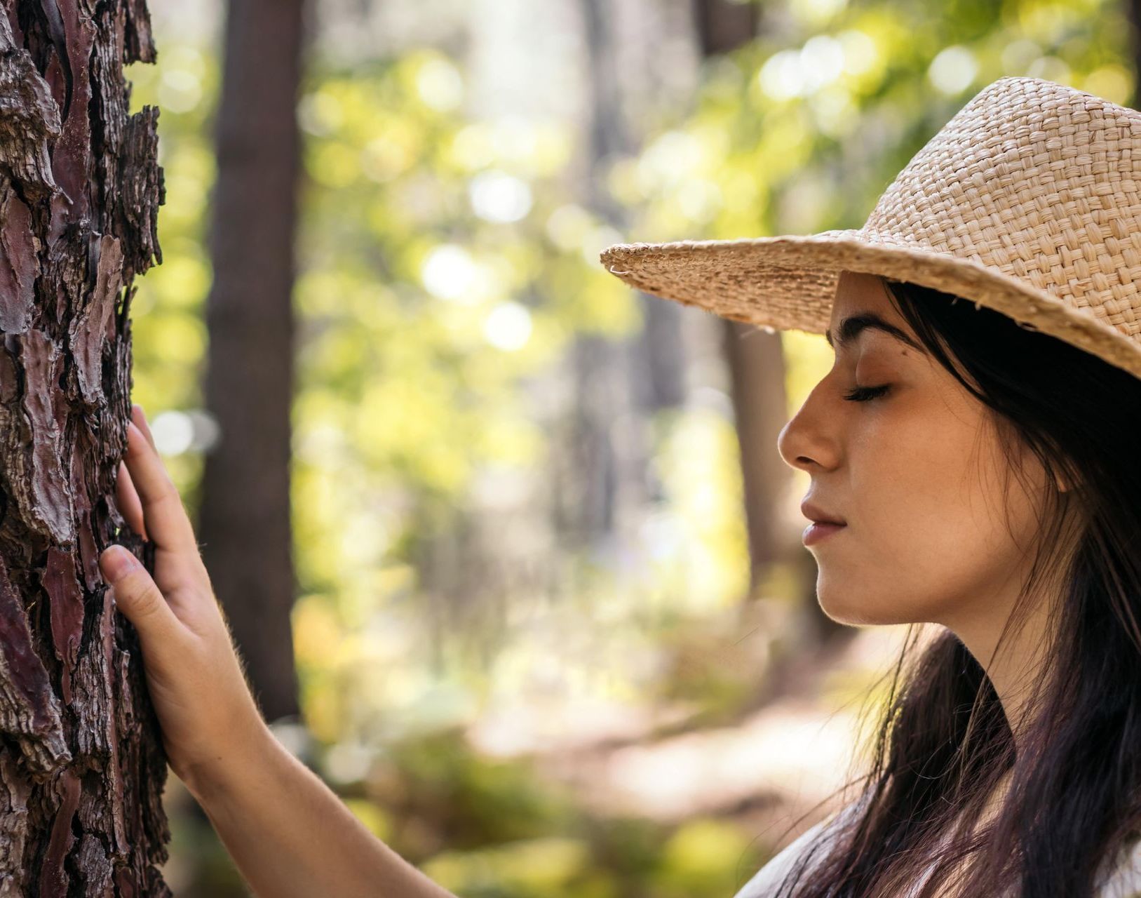 Woman in a straw hat touching a tree in a sunlit forest, eyes closed in contemplation.