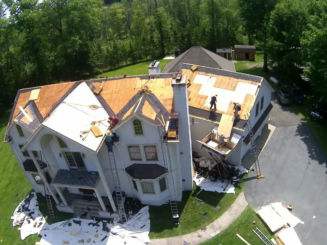 Roofers working on a white house, partially re-shingled. Green trees and lawn surround.