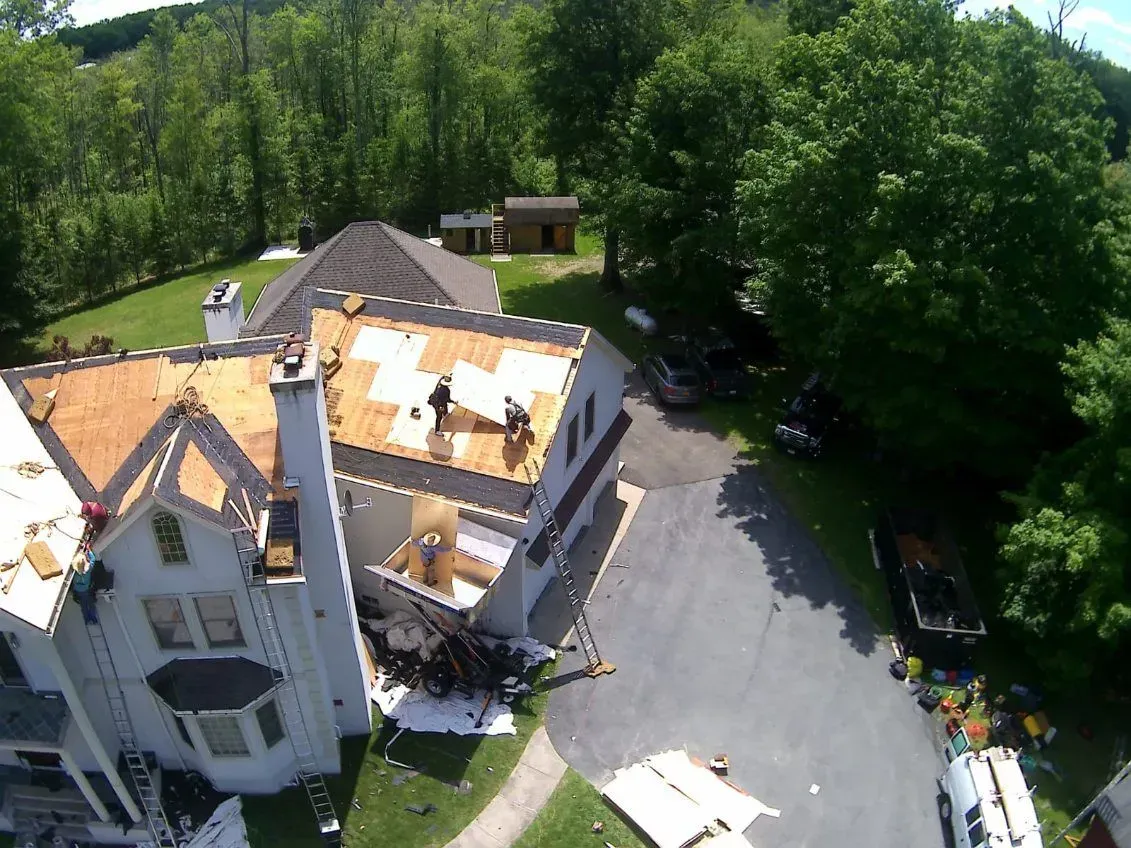Workers replacing a roof on a white house. Trees and parked cars surround the building.