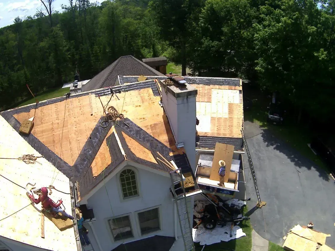 Roofing workers on a house. Wooden roof partially covered, workers on platforms. Green trees and driveway in the background.