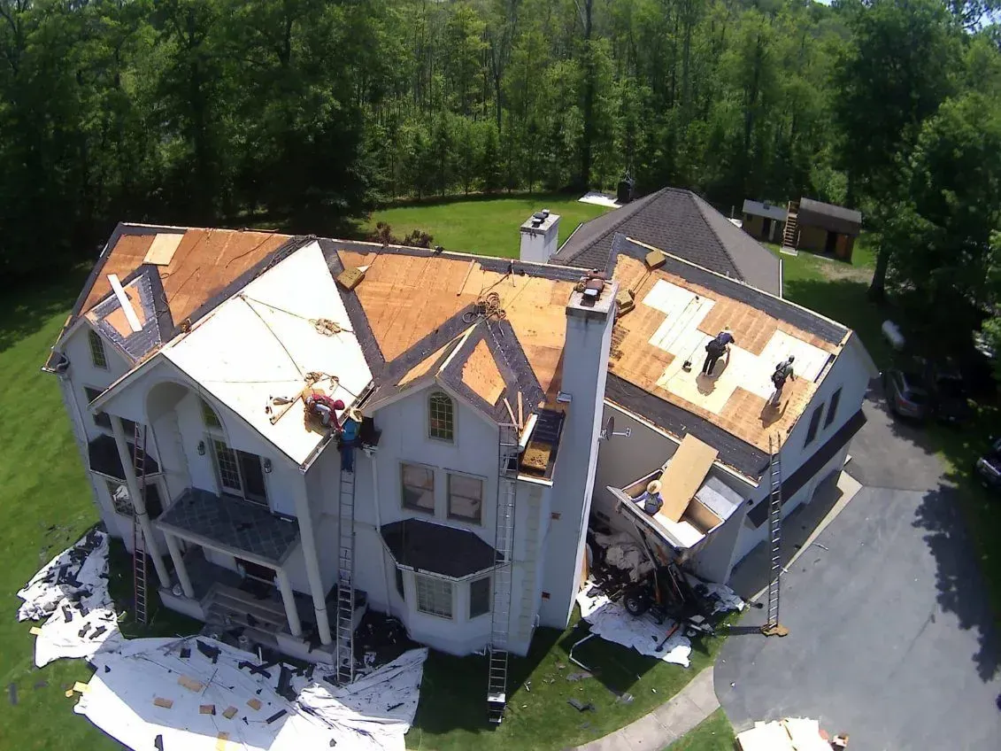 Roofing work in progress on a large, multi-story house. White tarps cover the yard, workers on the roof.