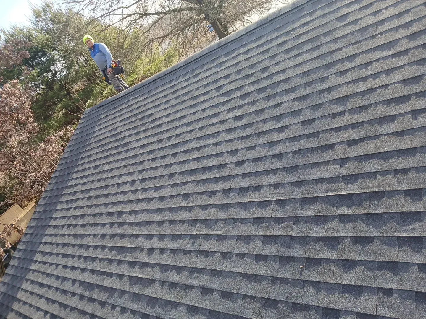 Roofer on a dark gray shingle roof in a sunny outdoor setting.