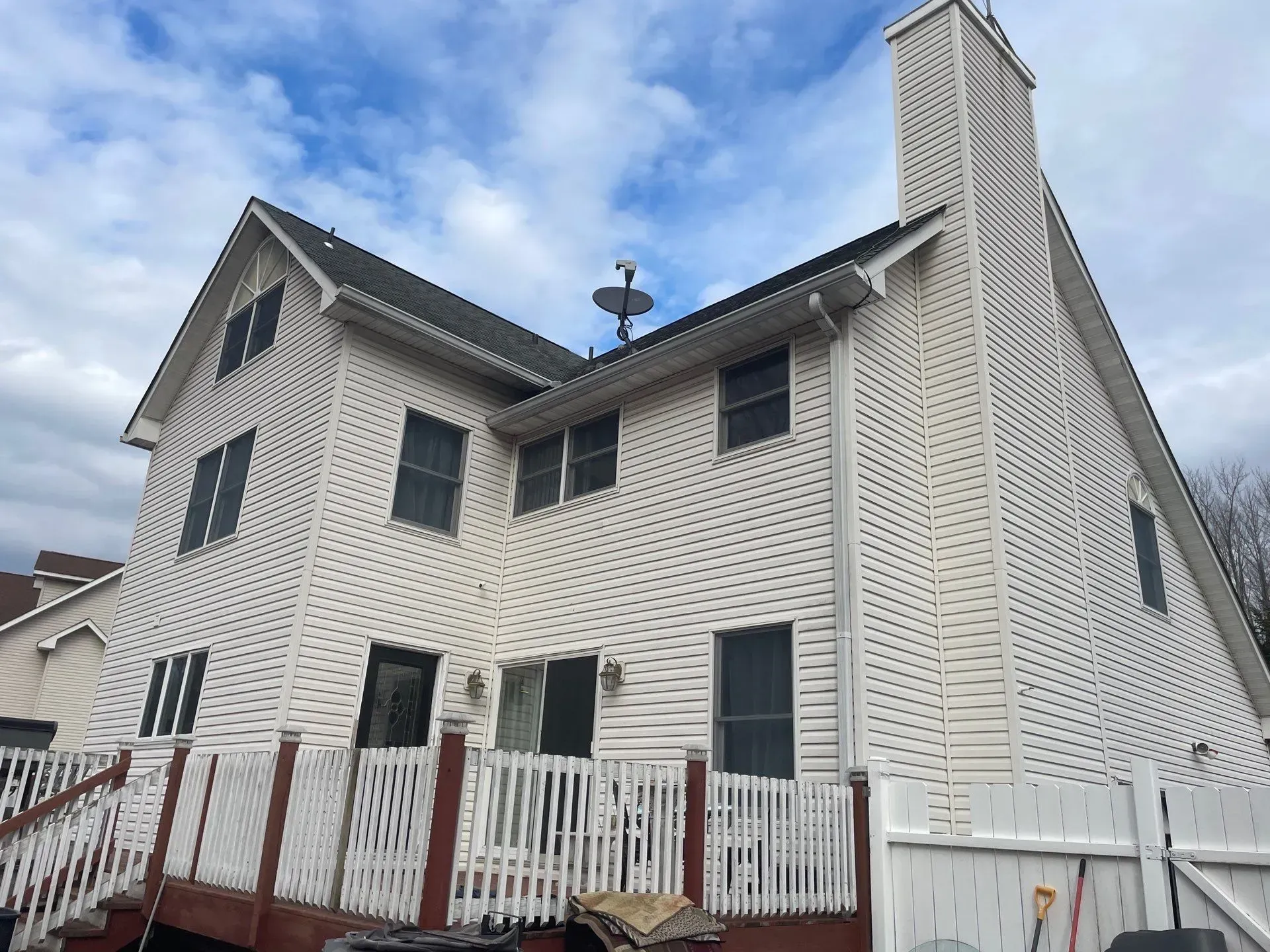 Two-story house with white siding, dark roof, chimney, and a deck with red trim, under a cloudy sky.