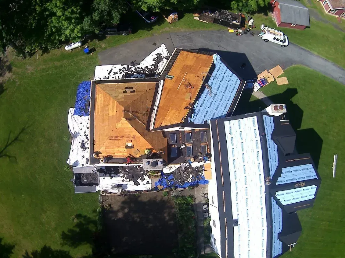 Aerial view of a house roof partially covered with blue tarps and new roofing materials. Green lawn surrounds it.