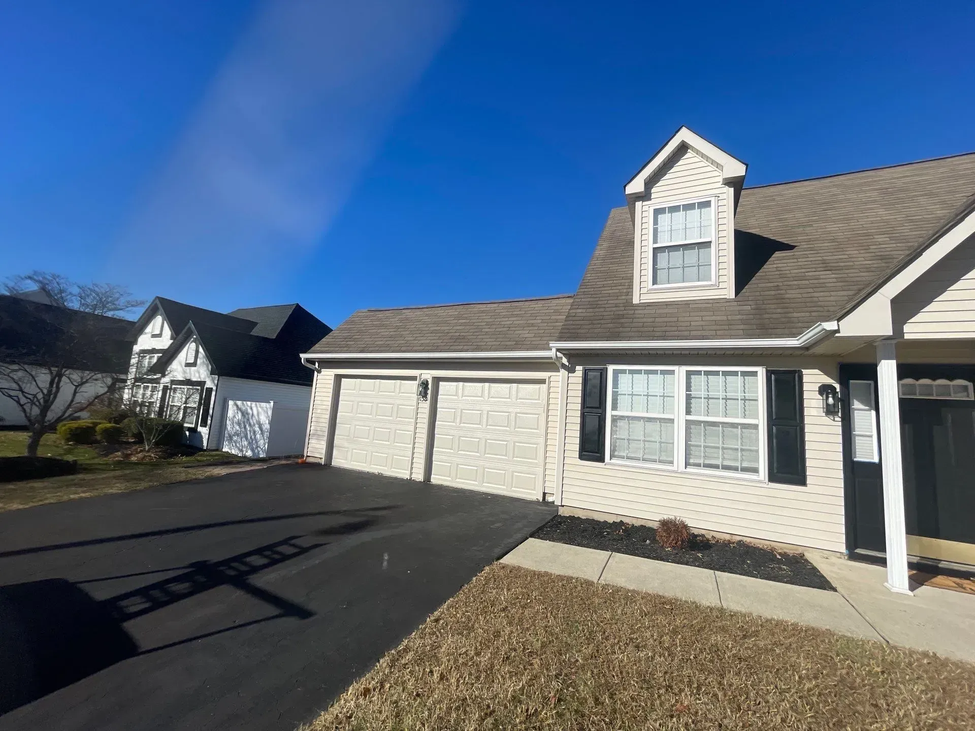 Tan house with three-car garage, dormer, and black shutters, set against a bright blue sky.