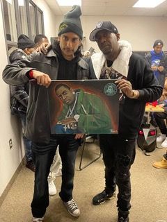 Two men are standing next to each other holding a painting of a man.