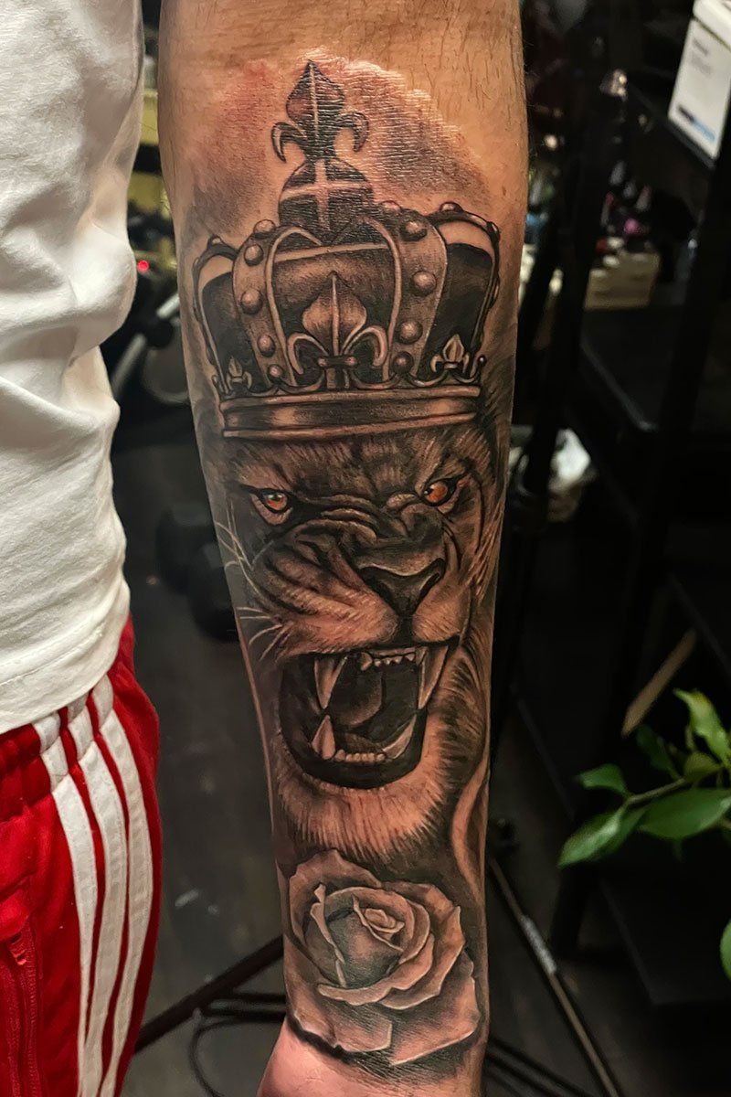 A man has a tattoo of a lion with a crown and a rose on his forearm.