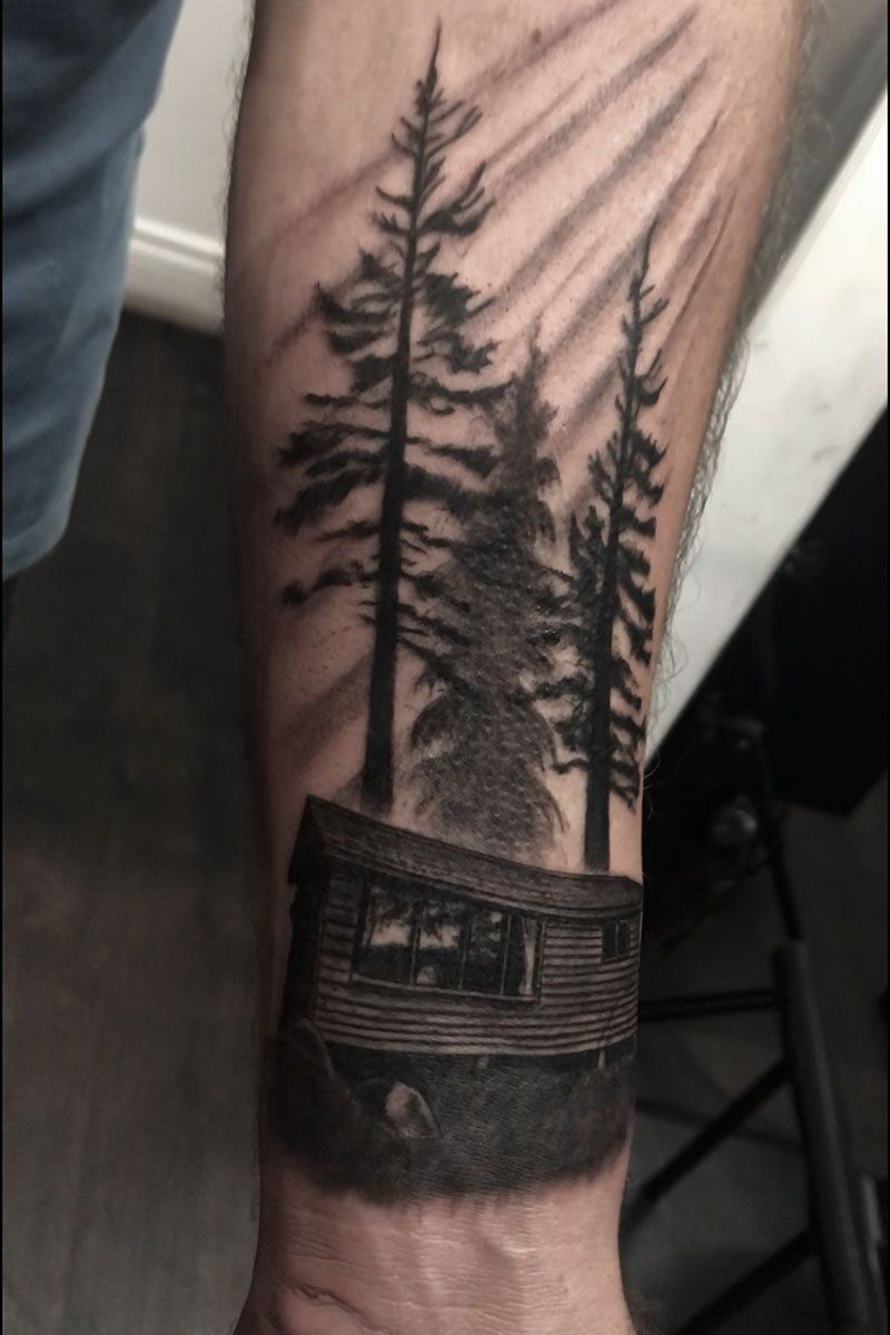 A man has a tattoo of a cabin in the woods on his forearm.