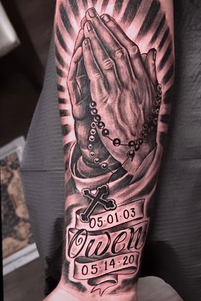 A man has a tattoo of praying hands with a rosary on his forearm.