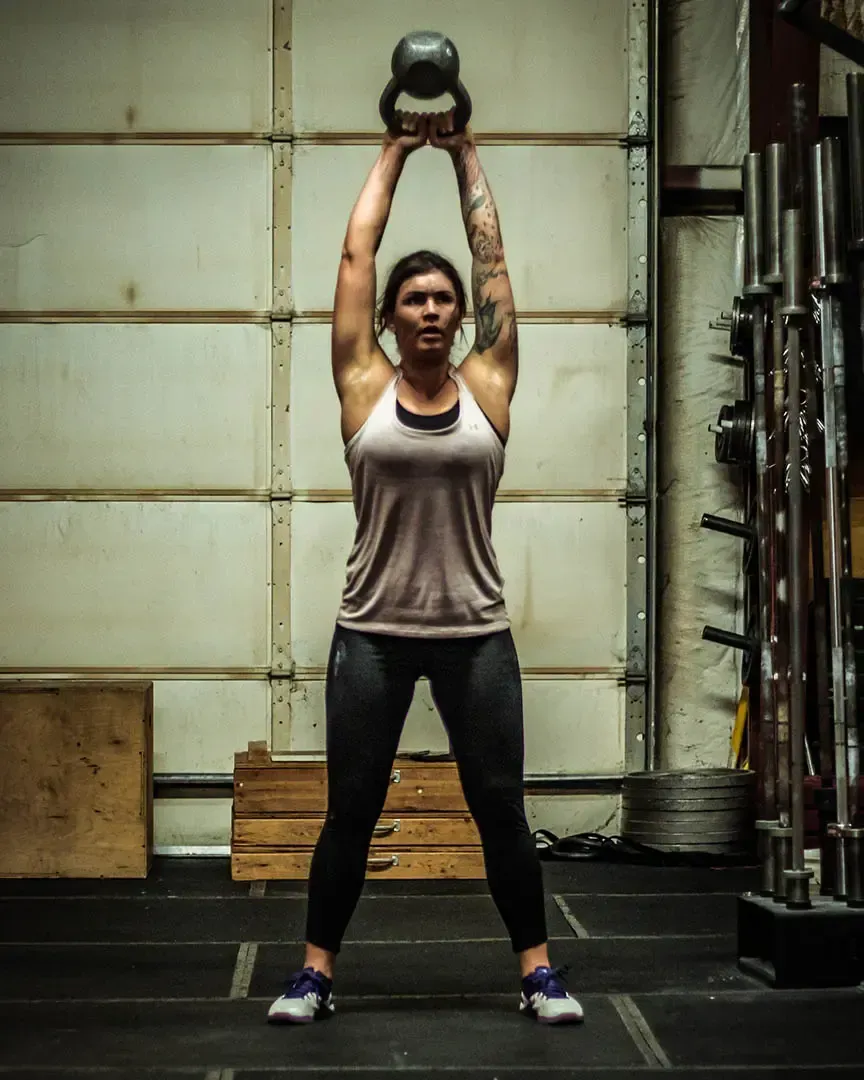 Woman in gym lifts kettlebell overhead, legs apart.